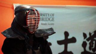 A figure dressed in kendo armour stands in front of a University of Cambridge banner