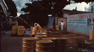 Imagem mostra toneis posicionados em frente a ferro-velho contaminado em acidente radiotivo em 1987, no Brasil, e homens trabalhando no processo de descontaminação no local