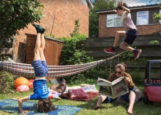 Frau liest Papier im Garten, während zwei Kinder um sie herum spielen