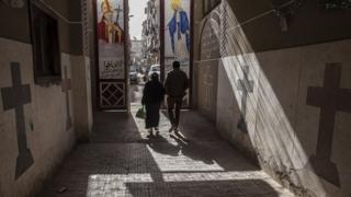 a man and woman walking through a sun-lit street with crosses and Christian motifs on the walls, file pic