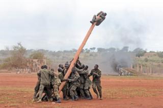 Soldiers hold on to one end of a large pole as their comrade clings to the other end in the air