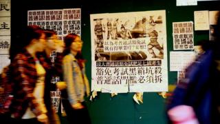 香港浸會大學逸夫校園內一群女學生走過貼上了反對普通話畢業門檻豁免試標語的學生會民主牆(BBC中文網圖片22/1/2018)