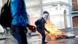 Dos personas protestan en Ecuador y de fondo se ve un colchón en llamas