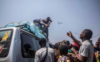 A man, standing on top of a car, throws a bottle of water to another man