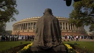 Статуя Махатмы Ганди возвышается над зданием индийского парламента, в то время как законодатели от оппозиционных партий образуют человеческую цепь в знак протеста против демонетизации правительством дорогостоящих законопроектов в Нью-Дели, Индия, в среду, 23 ноября 2016 года.
