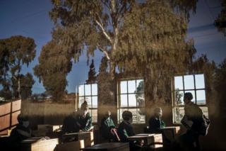 Secondary students sit in a classroom. The trees outside are reflected on the window pane.