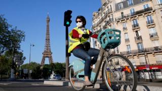 Una mujer con una máscara facial monta una bicicleta en París, la Torre Eiffel es capturada detrás de ella.