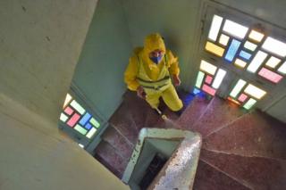 A man wearing a yellow PPE suit sprays mosque stairwell that is illuminated by coloured glass windows.