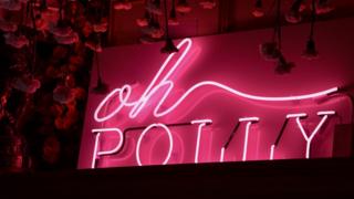 Розовый флуоресцентный знак логотипа Oh Polly на черном фоне.