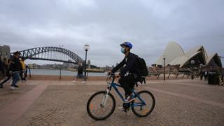 Мужчина носит маску для лица в качестве профилактической меры против коронавируса, когда он проезжает на велосипеде мимо моста Харбор-Бридж и Сиднейского оперного театра.