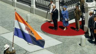 El expresidente sudafricano Pieter Willem Botha y su esposa Elize se paran frente al edificio del Parlamento en Ciudad del Cabo durante la apertura estatal del Parlamento en 1987. La bandera que flamea es la antigua insignia de la República de Sudáfrica, utilizada desde 1928-1994.