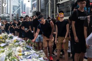 A line of people dressed in black flowers lay flowers on this site in a busy street of Hong Kong