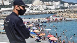 Ein Polizist, der aufgrund der COVID-19-Coronavirus-Pandemie in eine Maske gekleidet ist, beobachtet, wie sich Menschen am 15. August 2020 am Strand von El-Kettani im Vorort Bab el-Oued in Algeriens Hauptstadt Algier im Wasser abkühlen.