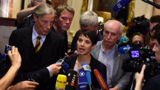 Frauke Petry with many microphones pointing at her, and supporters by her side