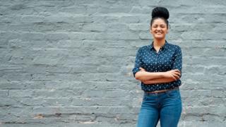 Mulher de camisa, calça-jeans, cabelos trançado preso no alto, sorrindo, com braços cruzados e em frente a uma parede
