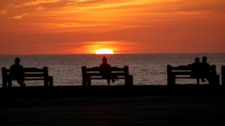 Two people sit on separate benches at sunset while a couple sit together on one bench