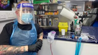 Tattoo artist Adam Grant wears a face mask, visor and disposable gloves and apron.