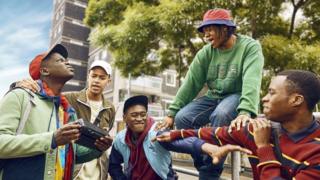 From left to right, the characters of Dane played by Yus Jamal Crookes - who is wearing looking up, wearing a red cap and green jacket, holding a black radio, Junior played by Gabriel Robinson - who is wearing a brown jacket and white cap with his arm around Dane's shoulder, Bishop played by Tienne Simon - who is wearing a blue jacet with dark blue sleeves and a maroon hoody underneath with a dark coloured cap, reaching out his left arm, Kai played by Shanu Hazzan - wearing a green top and blue jeans with a silver chain around his neck and pink and blue buckethat looking down at Tienne, and Bayo played by Juwon Adedokun, wearing a maroon sweater with blue and yellow lines, pointing towards Tienne. The background is of a large tower block covered by green trees.
