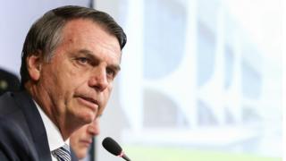 Bolsonaro fala no microfone em posição de perfil, com imagem da fachada do Palácio do Planalto ao fundo