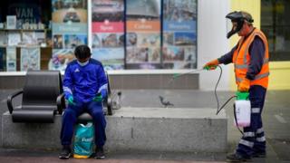 Council worker in Leicester spraying disinfectant on a public bench, close to someone sitting in a mask after government announces lockdown will continue in the city