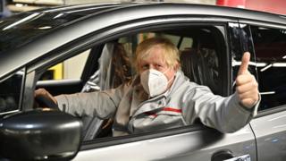 Boris Jonson in a silver car giving a thumbs up out the driver window. He is wearing a grey jacket and a medical face mask.
