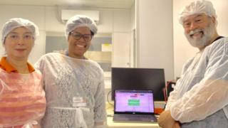Com jalecos e toucas, Claudia Gonçalves, Jaqueline Goes e Claudio Sacchi sorriem para foto de dentro de laboratório