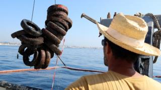 man in straw hat watching tyres being winched out of the sea by a boat