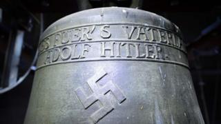 Una campana de la época nazi, que tiene grabada una esvástica, una línea que dice "Todo por la patria" y otra que dice "Adolf Hitler".