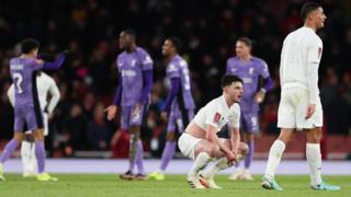 Declan Rice crouches down as Liverpool celebrate in the background
