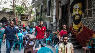 A cafe server carries hot coals for customers' waterpipes (shisha) at a cafe in downtown Cairo with a mural seen painted in the background (R) depicting the smiling face of Liverpool's Egyptian midfielder Mohamed Salah on 30 April 2018