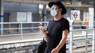 Man in a mask, sunglasses and a hat walks along a platform at a London Underground station with a phone in his hand