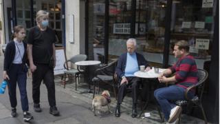 Padre con máscara e hija pasan junto a dos hombres sentados afuera de un café en Soho, Londres