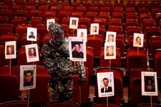 A performer in a metallic costume adjusts seat markings in a theatre