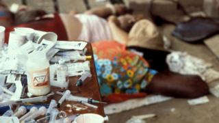 Photograph of medical byproducts and syringes conspicuous, with people lying visible in the foreground