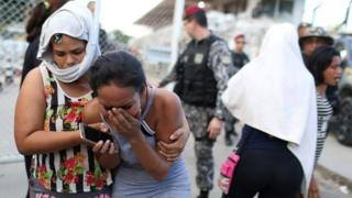 Mulheres choram e se consolam do lado de fora de presídio em Manaus, com agentes de segurança e cerca aparecendo ao fundo