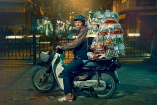 A man is posed on a motorcycle with an array of multicoloured fish in plastic bags hanging in rows on the back of the bike