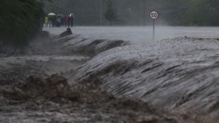 People watch as flash floods rage past a part of the main highway to Kenya"s border town with Tanzania on March 15, 2018 in Isinya, Kajiado county, around 58 kilometres (36 miles) southeast of capital Nairobi.