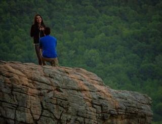 Дэвид стоит на одном колене, а Дженни улыбается, сидя на скале Хоксбилл