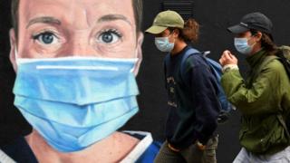Pedestrians wearing a face mask or covering due to the COVID-19 pandemic, walk past graffiti by the artist @akse_p19, depicting a nurse in scrubs and a face mask, but with an Angel's halo above her head, in Manchester, northwest England on August 3, 2020