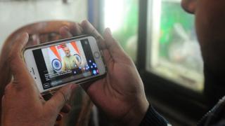 Индийский лавочник использует свой мобильный телефон, чтобы посмотреть прямую трансляцию премьер-министра Нарендры Моди в Аллахабаде 31 декабря 2016 года.