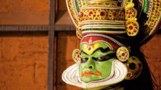 Un actor con la cara pintada con maquillaje verde y sombras negras alrededor de los ojos y en la frente. También tiene un elaborado sombrero ornamental.