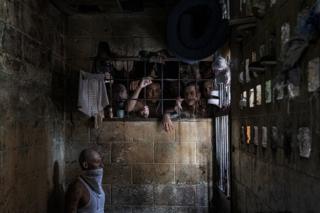 Inmates look out of an overcrowded cell, while a man wears a bandana across his face because of the putrid smell. Penal Center of Quezaltepeque, El Salvador. November 9, 2018.