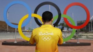 Homem com camiseta do RIo2016 tira foto diante de arcos olímpicos
