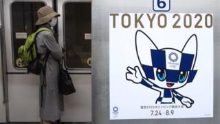 Пассажир в маске стоит рядом с плакатом с олимпийским талисманом Токио-2020 Мираитовой в поезде в Токио 20 апреля 2020 года.