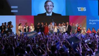 No palco, Alberto Fernandez e a ex-presidente Cristina Kirchner comemoram os resultados das eleições em Buenos Aires, saudados por dezenas de pessoas segurando bandeiras da Argentina no público