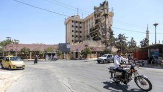 Мужчина проезжает на мотоцикле по улице мимо разрушенного бывшего здания отеля Carlton, которое служит больницей Сирийского Красного Полумесяца, в удерживаемом повстанцами северном сирийском городе Идлиб