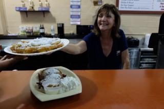 A woman behind a counter hands over a funnel cake in a paper plate