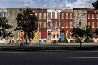 Young boys ride a bicycle in front of a row of lined up and abandoned houses in Baltimore.