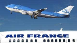 The top of a plane, sporting the AIR FRANCE logo, occupies the bottom third of this photo, while a blue KLM plane hovers over it.