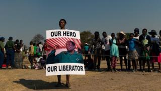 A young woman holds a placard reading 'Our hero, our liberator' villager queue to view the coffin of late former Zimbabwean president Robert Mugabe lying in state at Murombedzi Growth Point, about 107 km northwest of Harare, Zimbabwe, on September 16, 2019, as people have been accorded the opportunity to view Mugabe's body a week after his death. - The remains of former Zimbabwe president Robert Mugabe were taken to his village for a wake on September 16, a family member said, as his final burial is prepared in about a month. Mugabe died a week ago aged 95 in Singapore, nearly two years after he was ousted in a 2017 coup that ended nearly four decades of increasingly autocratic rule. After a state funeral on September 14 in the capital Harare attended by African leaders, his body went to his rural village of Kutama, 90 kilometres (55 miles) to the west, to allow villagers to pay tribute and bid farewell.